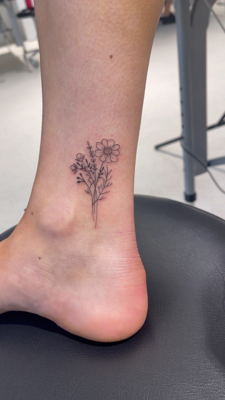 Small black ink daisy and wildflower tattoo on a person's ankle