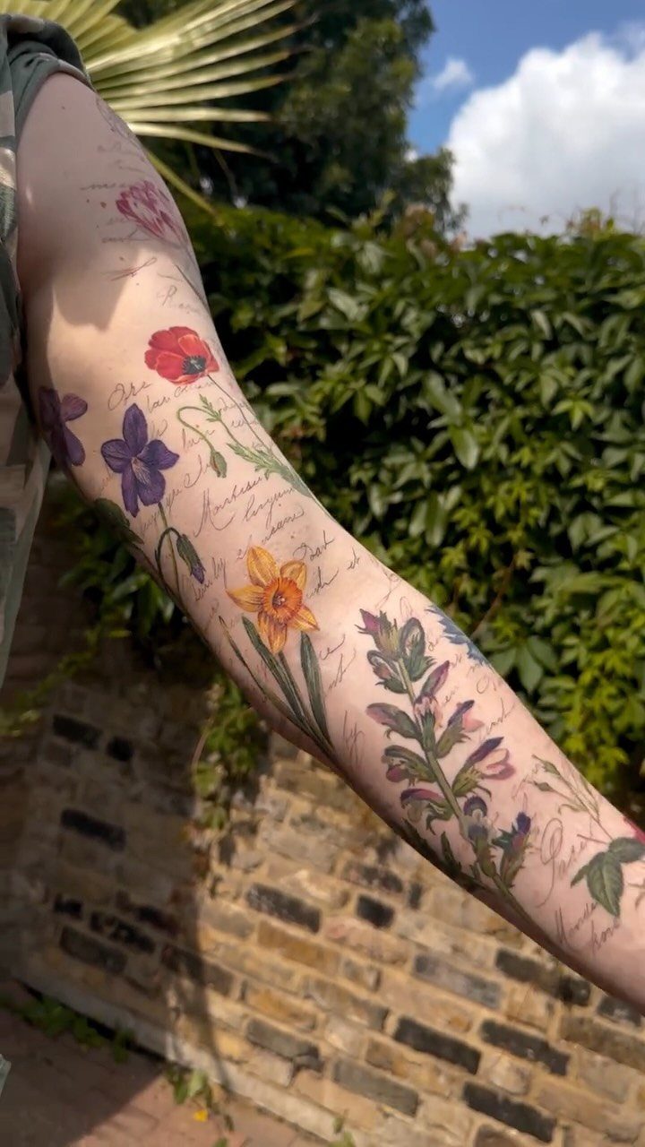 Forearm tattoos of colorful flowers and cursive script text against a garden background.