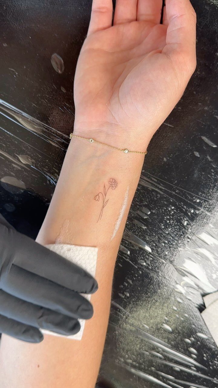 Small snowdrop flower tattoo on a person's inner forearm with a gold bracelet.