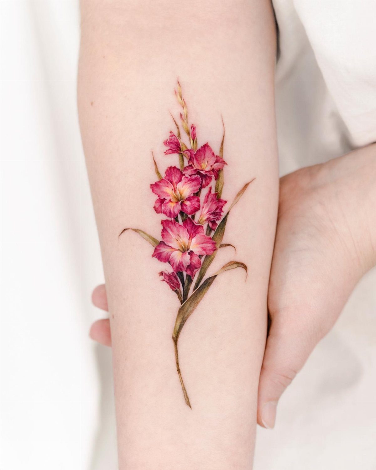 Pink gladiolus flower tattoo with green leaves on a person's inner forearm