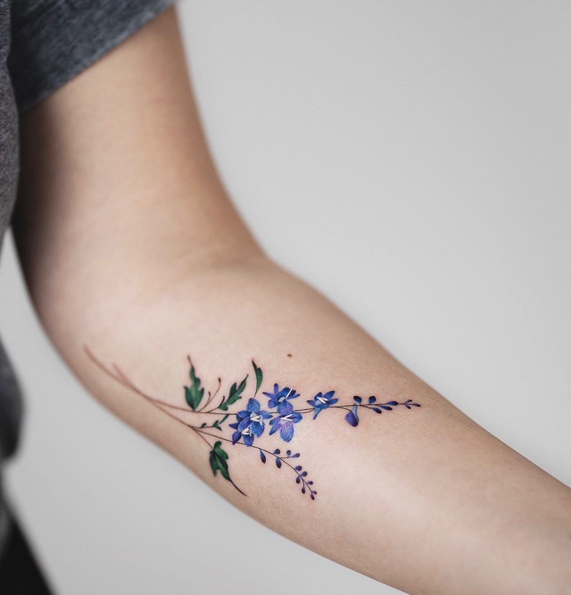 Blue larkspur flower tattoo with green leaves on a person's inner forearm.