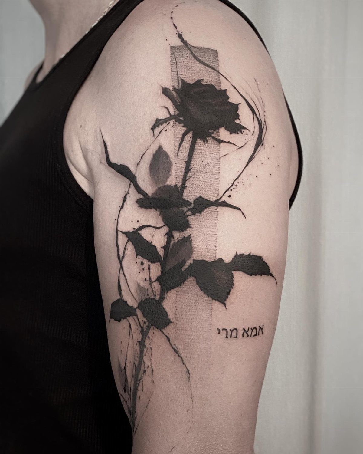 Black ink rose tattoo with splattered effects and Hebrew text on an upper arm.