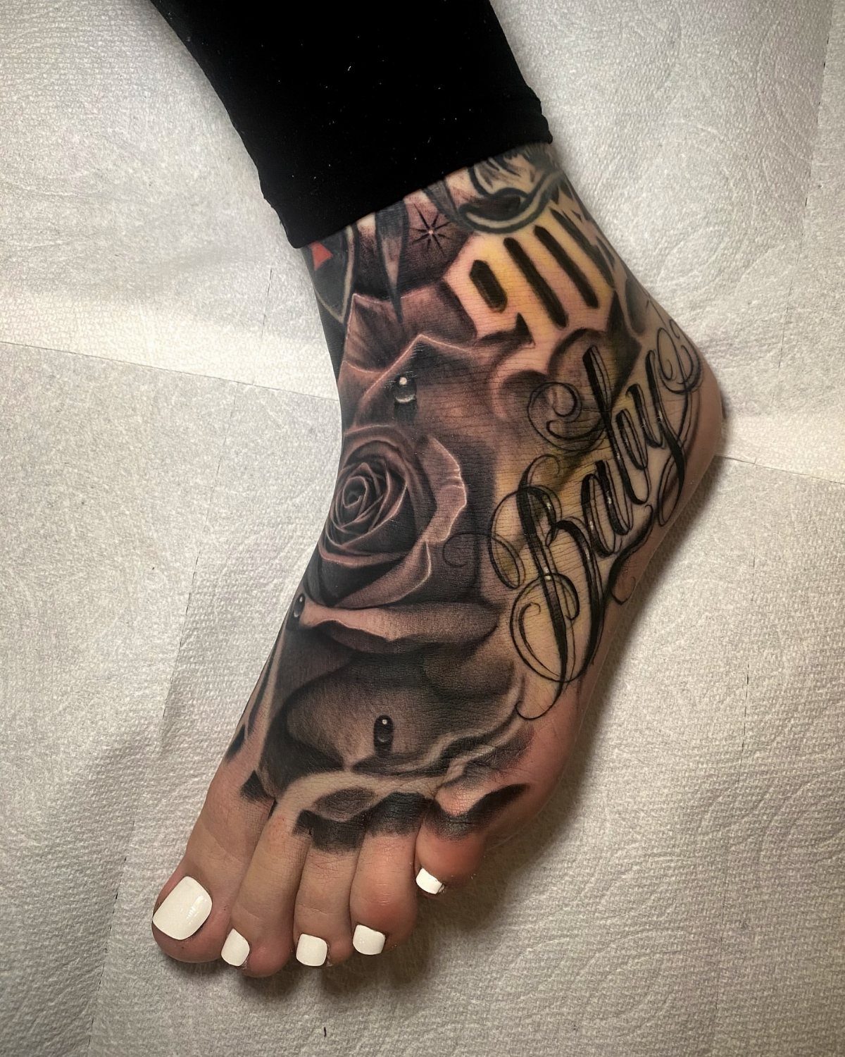 Detailed black and gray rose tattoo with script on a person's foot.