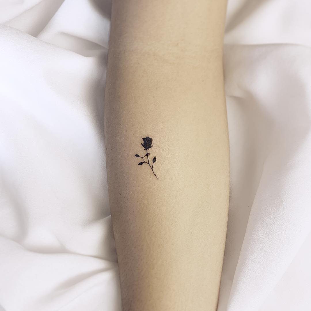 Small black rose tattoo with stem and leaves on a pale forearm.