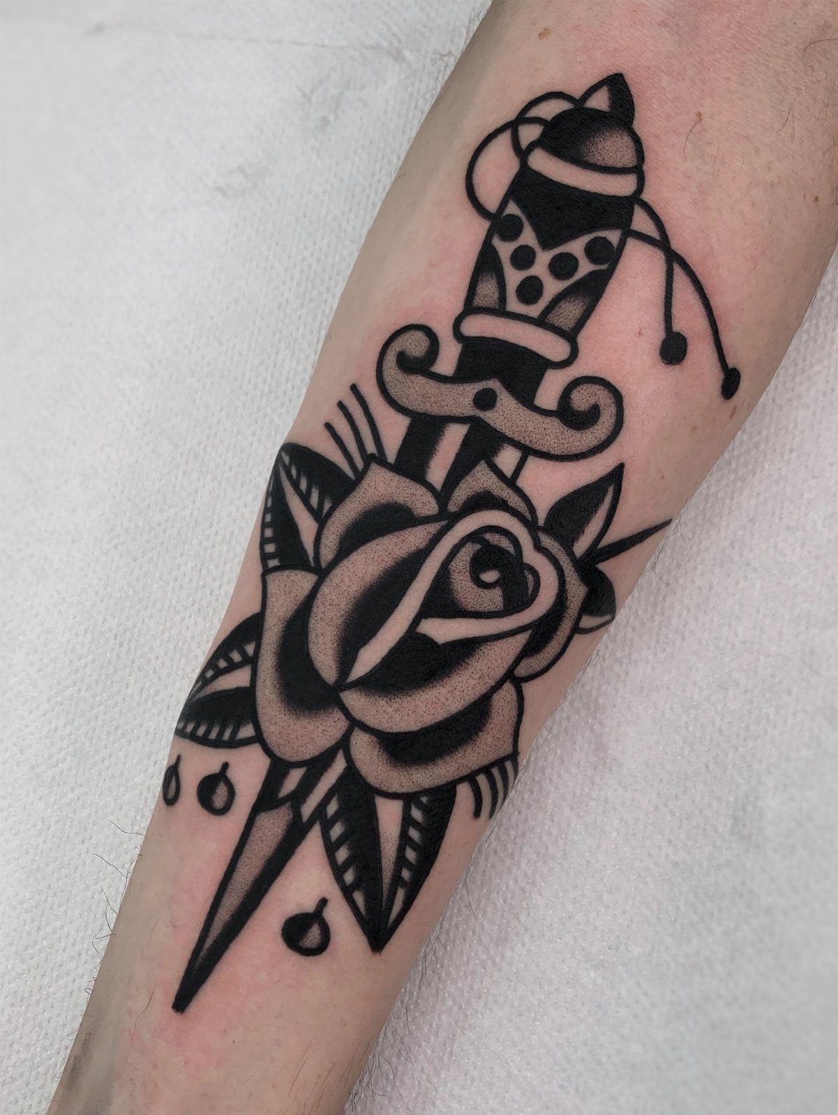 Black ink tattoo of a rose and dagger on a person's forearm
