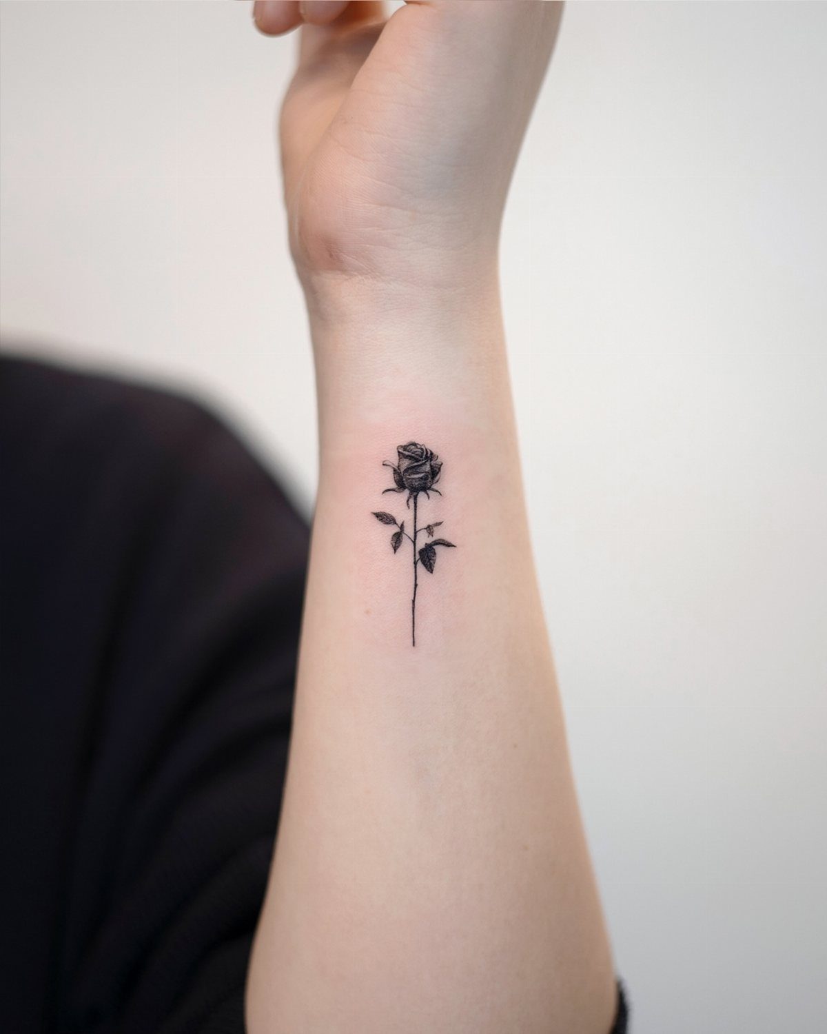 Small black rose tattoo with stem and leaves on a person's inner forearm.