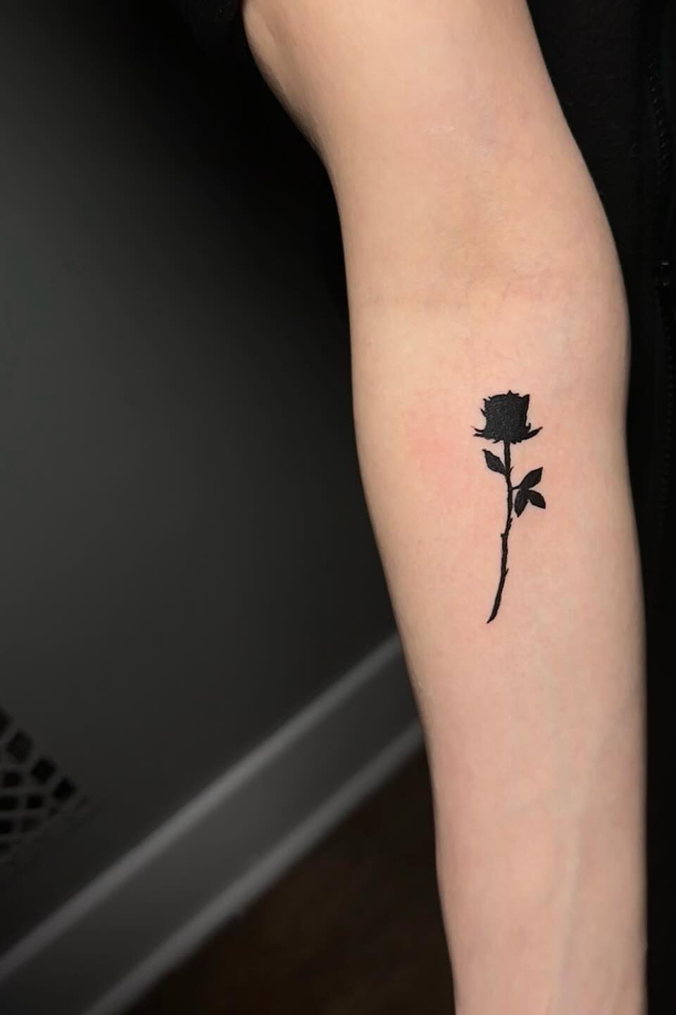 Small black rose silhouette tattoo on a person's inner forearm.