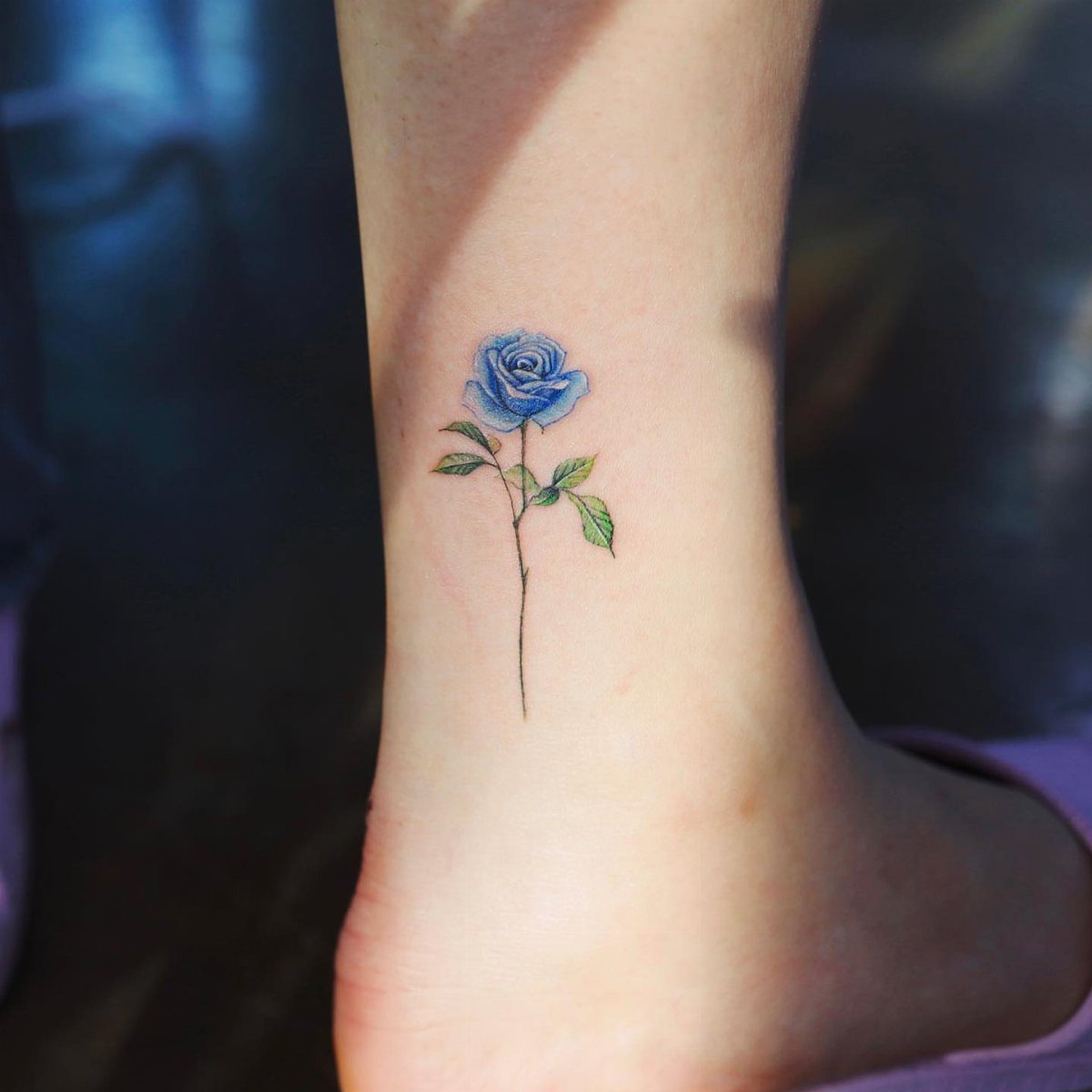 Blue rose tattoo with green leaves on a person's inner ankle