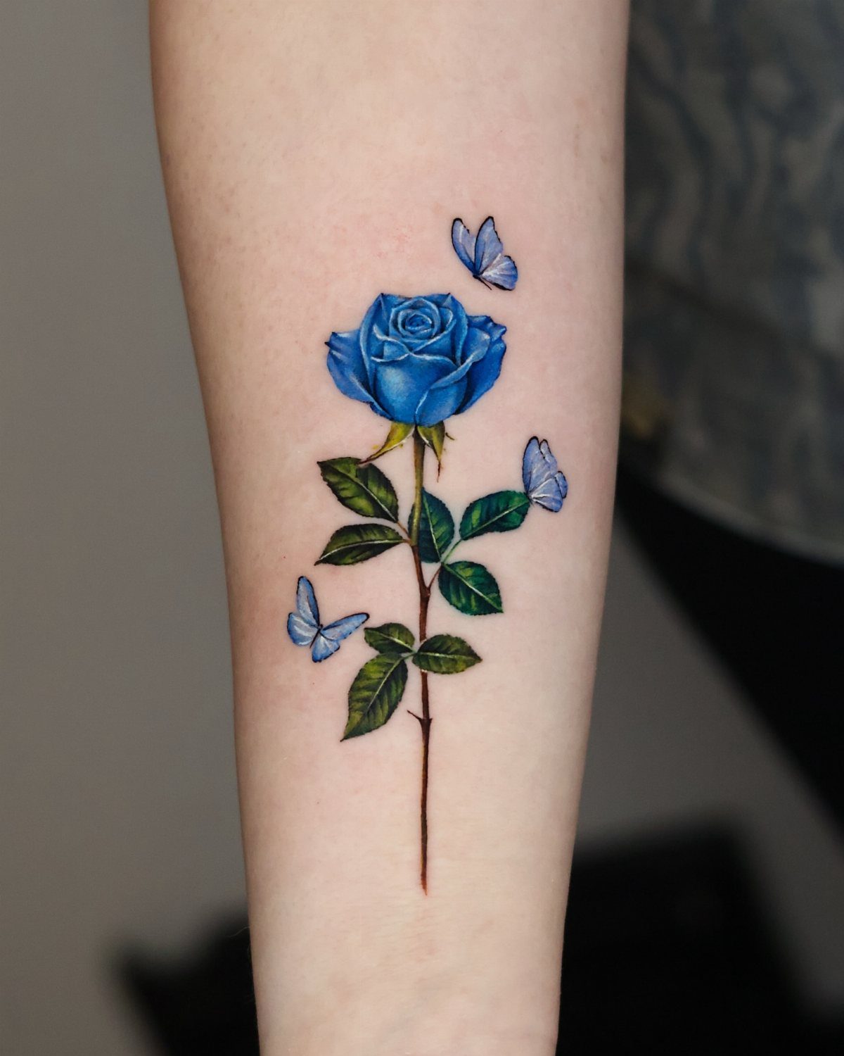 Blue rose tattoo with green leaves and blue butterflies on a forearm