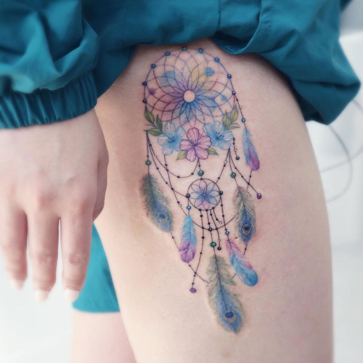 Dreamcatcher tattoo with blue flowers and purple feathers on a person's thigh