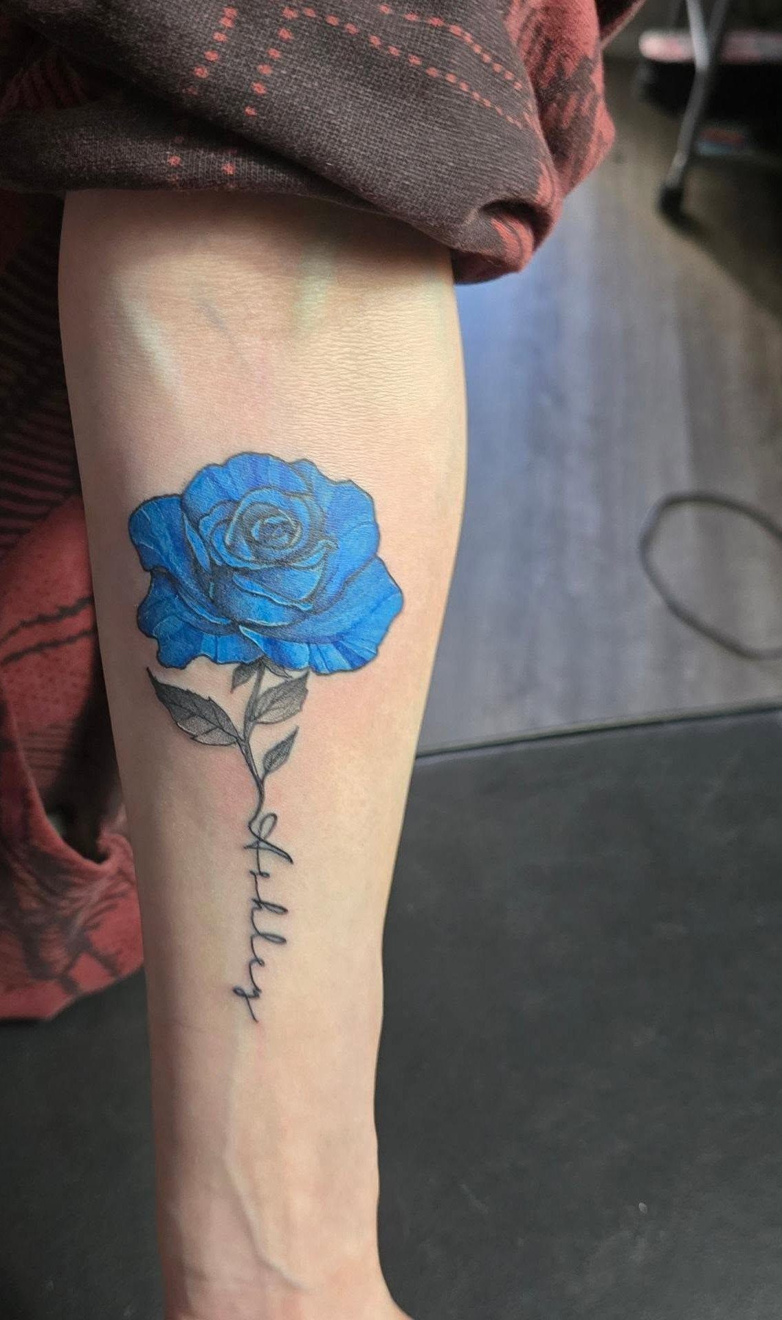 Blue rose tattoo on a lower leg with the name Kylie written below.