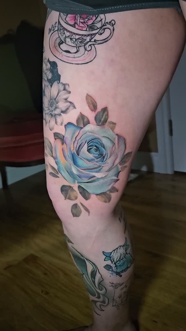 Blue rose tattoo with green leaves on a person's thigh and leg