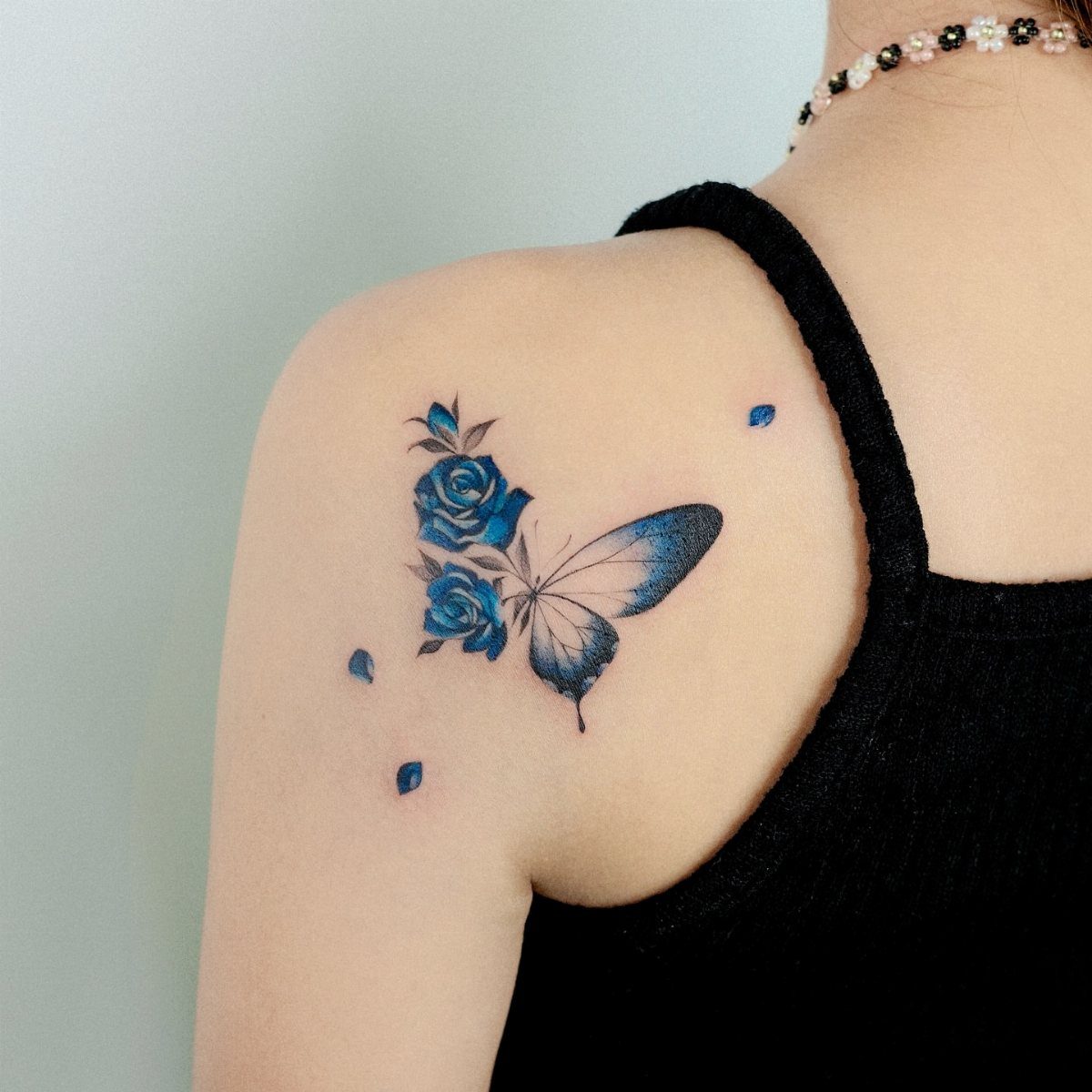 Blue rose and butterfly wing tattoo on a person's shoulder with floating petals