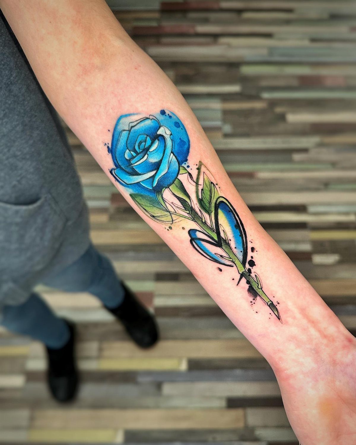 Blue rose tattoo with green leaves and a small heart on a forearm