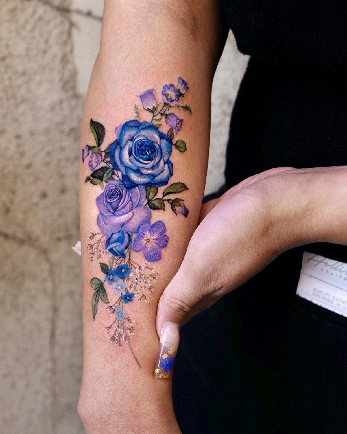Blue and purple rose tattoo with smaller flowers on a person's forearm.