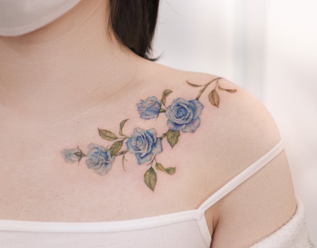 Blue rose tattoo with green leaves along the collarbone and shoulder of a person