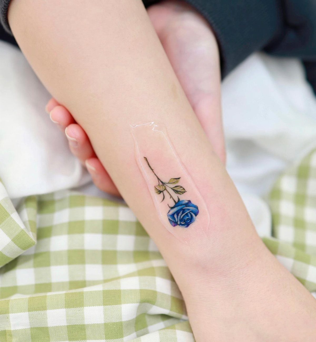 Small blue rose tattoo with green leaves on a forearm under clear bandage