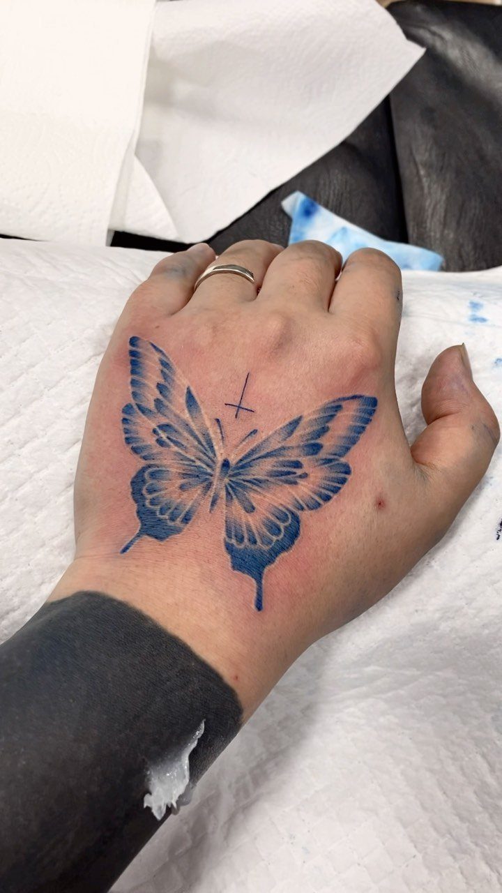 Blue butterfly tattoo on the back of a hand with a small cross.