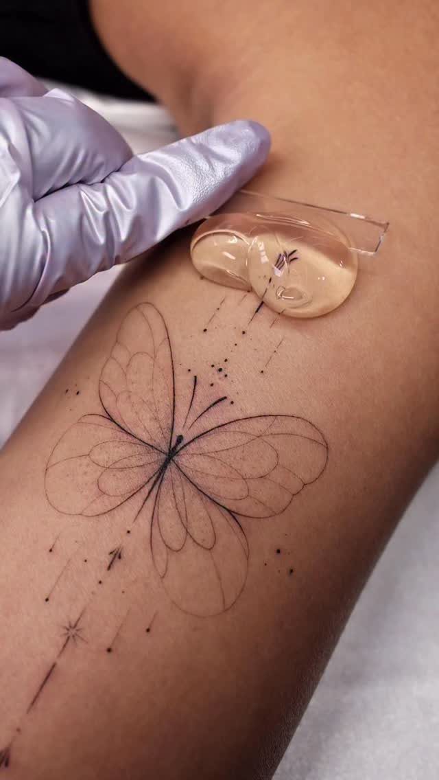 Fine line butterfly tattoo on an arm with a gloved hand nearby.