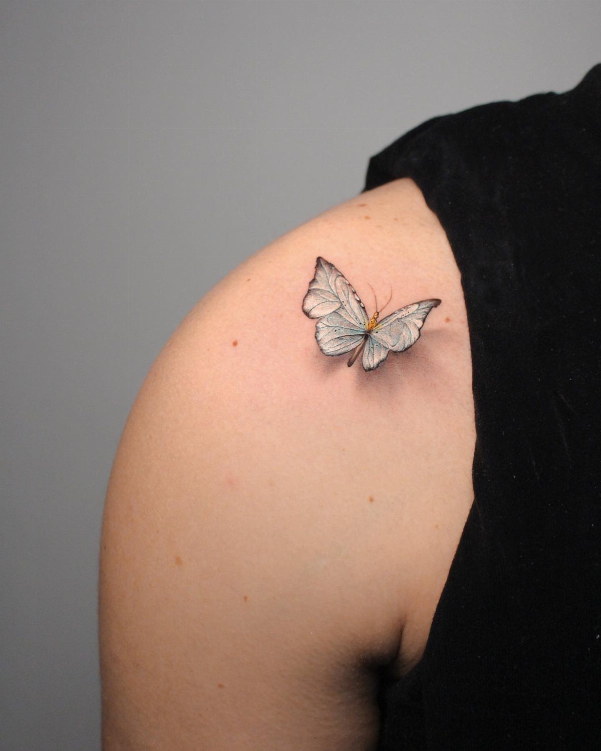 Small realistic white butterfly tattoo on a person's shoulder with detailed wing shading.