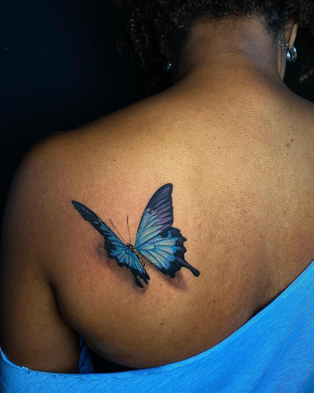 Blue and purple butterfly tattoo realistically positioned on a person's shoulder.