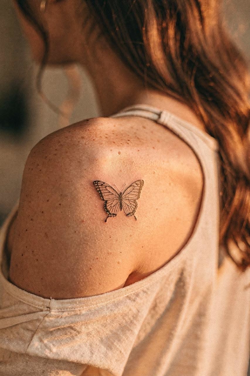 Small detailed butterfly tattoo on a freckled shoulder of a woman wearing white.