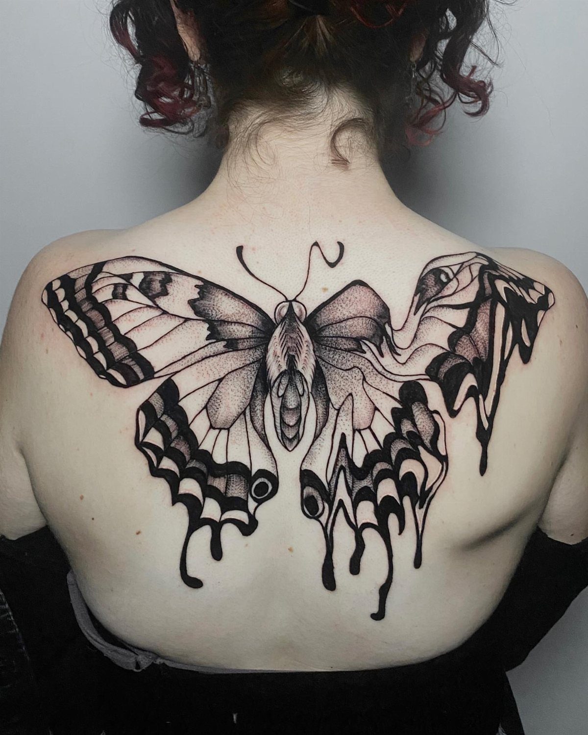Large black ink butterfly tattoo with dripping edges on a person's upper back