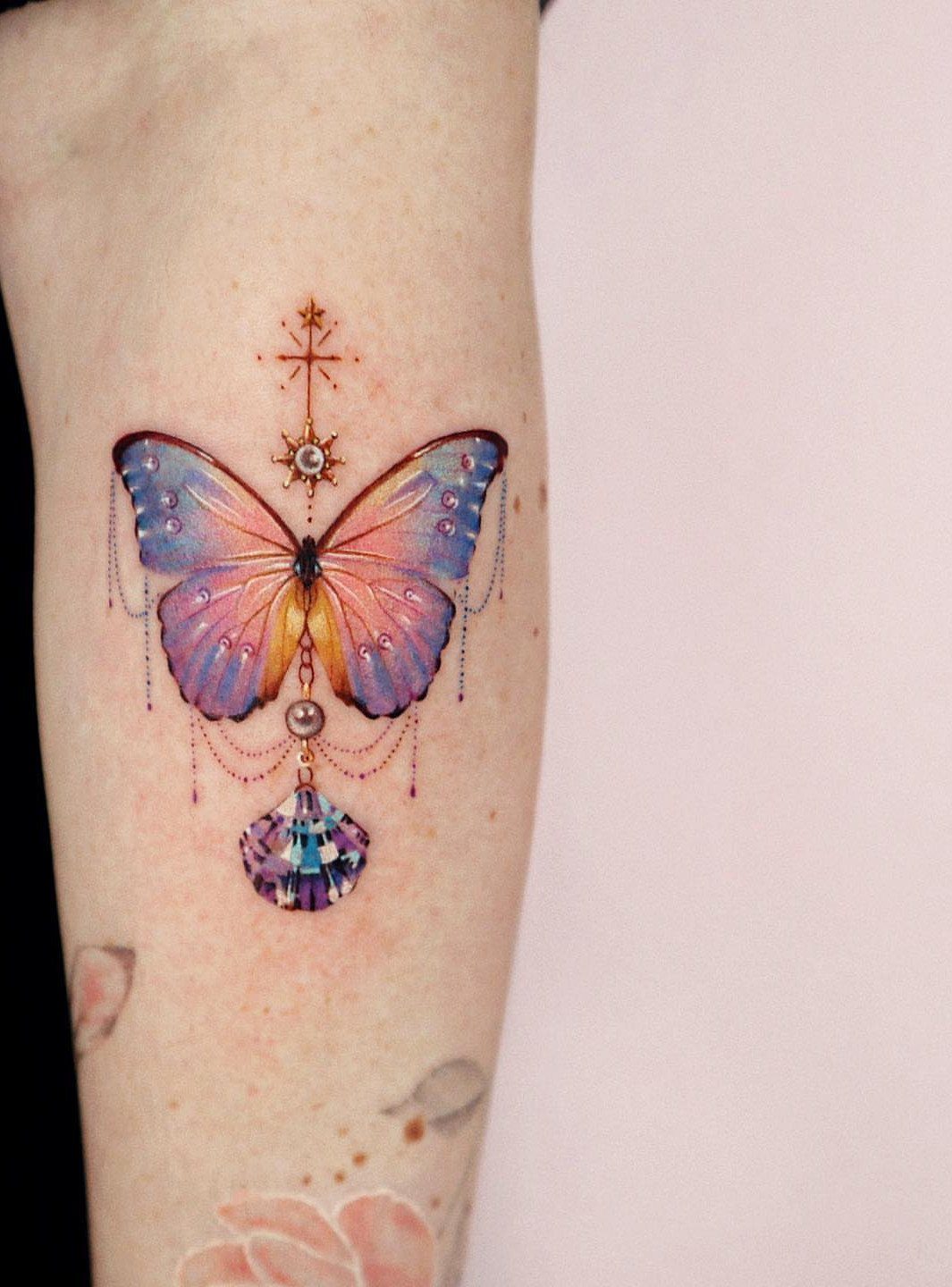 Butterfly tattoo with purple and blue wings, a golden cross, and a gemstone.