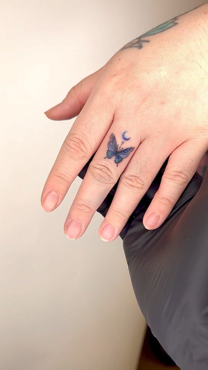 Small blue butterfly and crescent moon tattoo on a person's middle finger