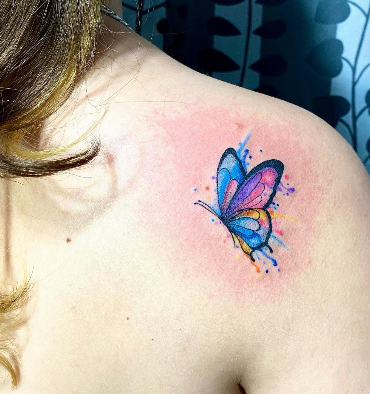 Watercolor butterfly tattoo with blue and purple splatters on a woman's shoulder
