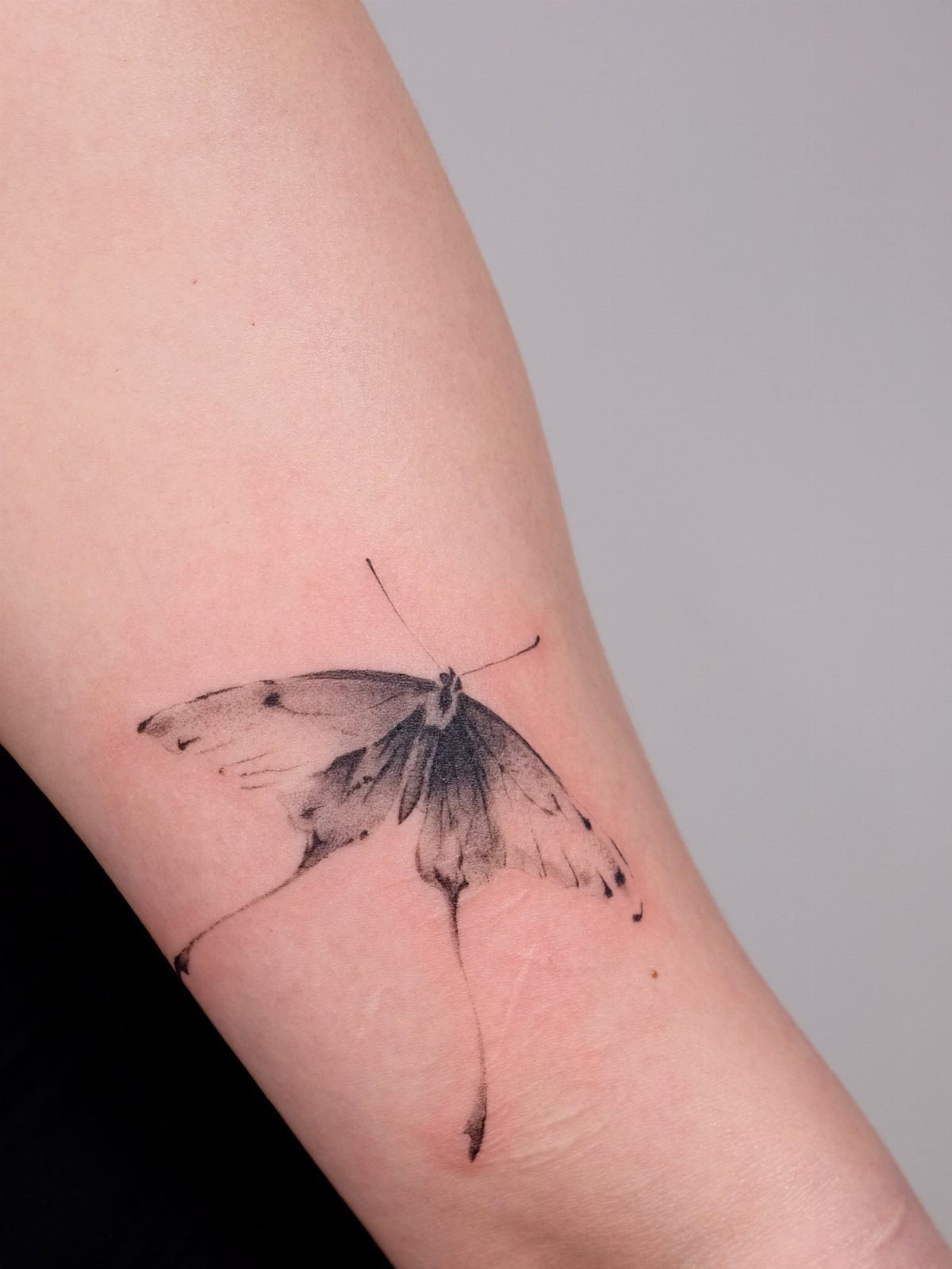 Butterfly tattoo on a forearm with soft, fading black ink wing details.