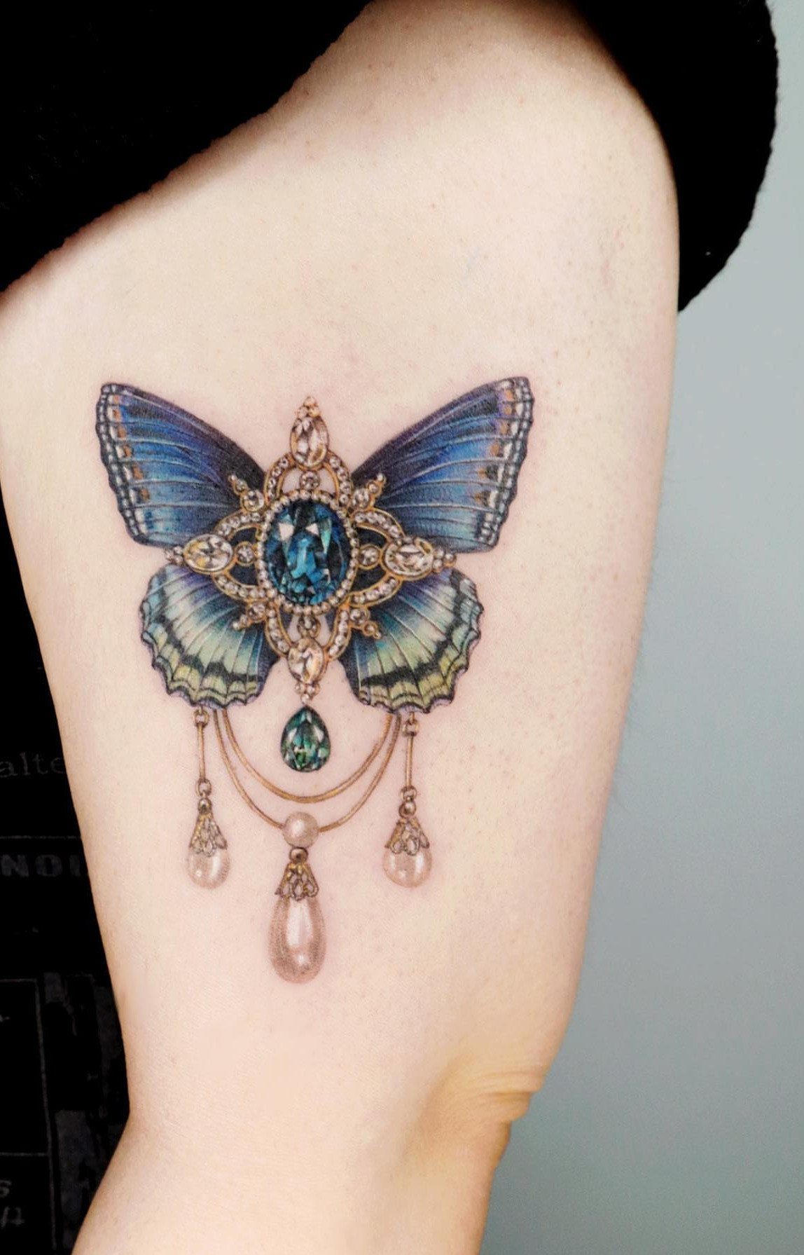 Butterfly tattoo with blue jewel-tone wings and ornate gold jewelry on an arm