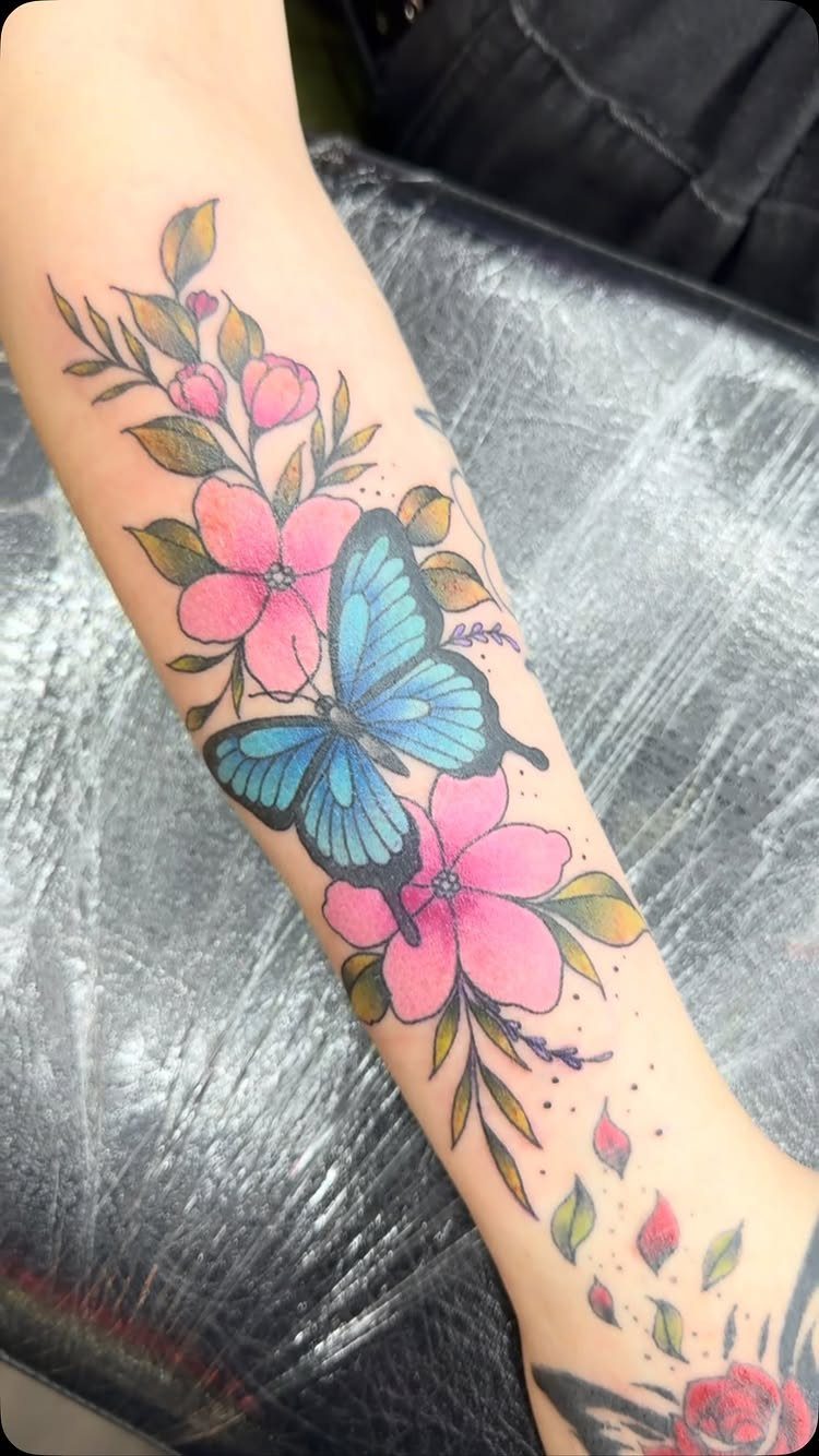 Blue butterfly tattoo surrounded by pink flowers and green leaves on a forearm