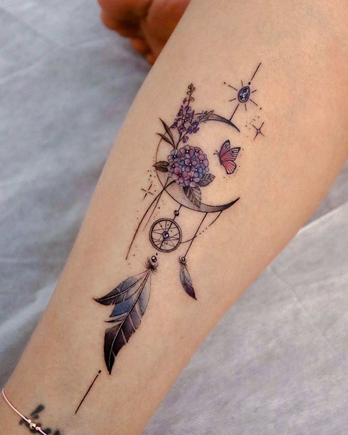 Colorful tattoo on an arm featuring a butterfly, dreamcatcher, moon, and purple flowers.