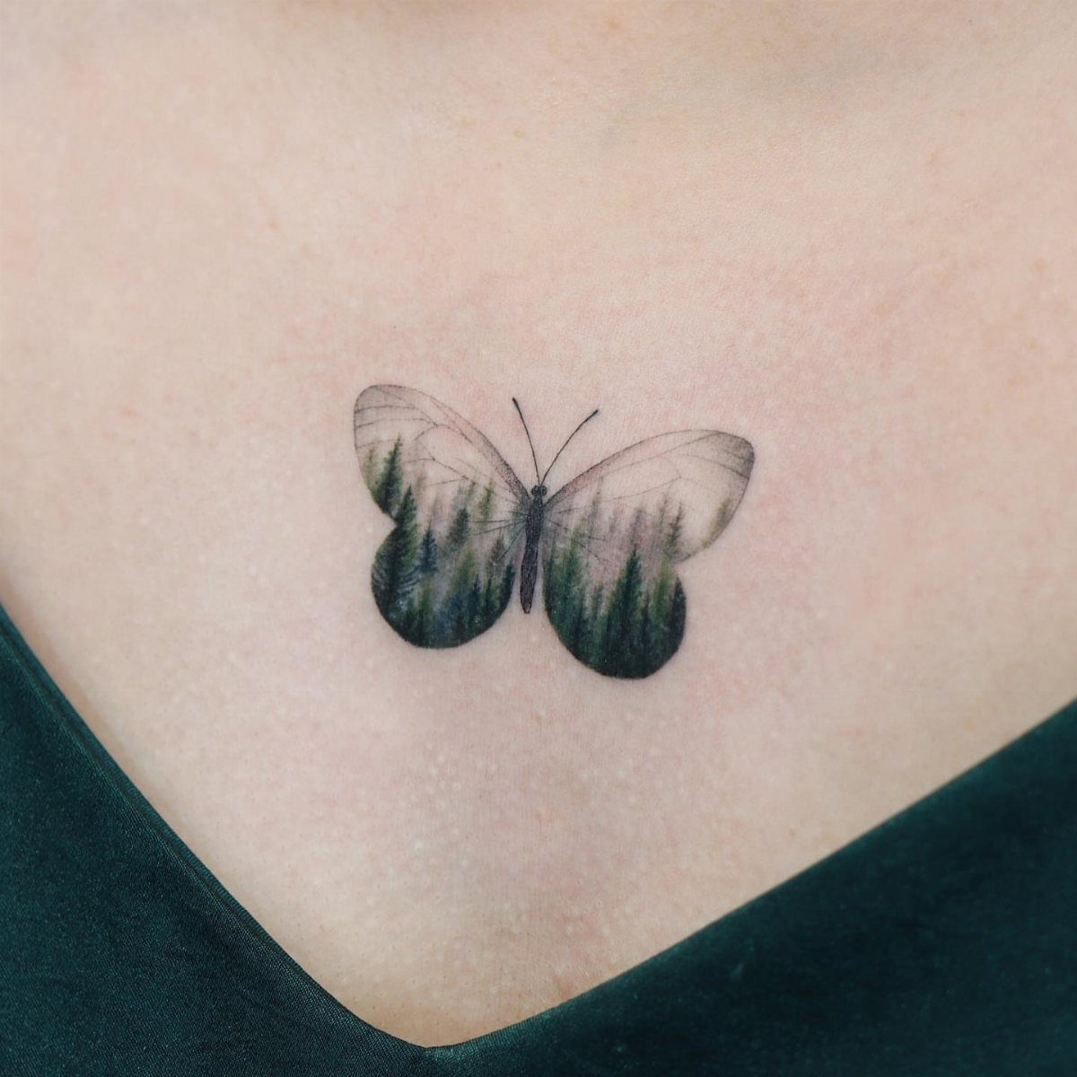 Butterfly tattoo on chest featuring a green pine forest landscape inside the wings