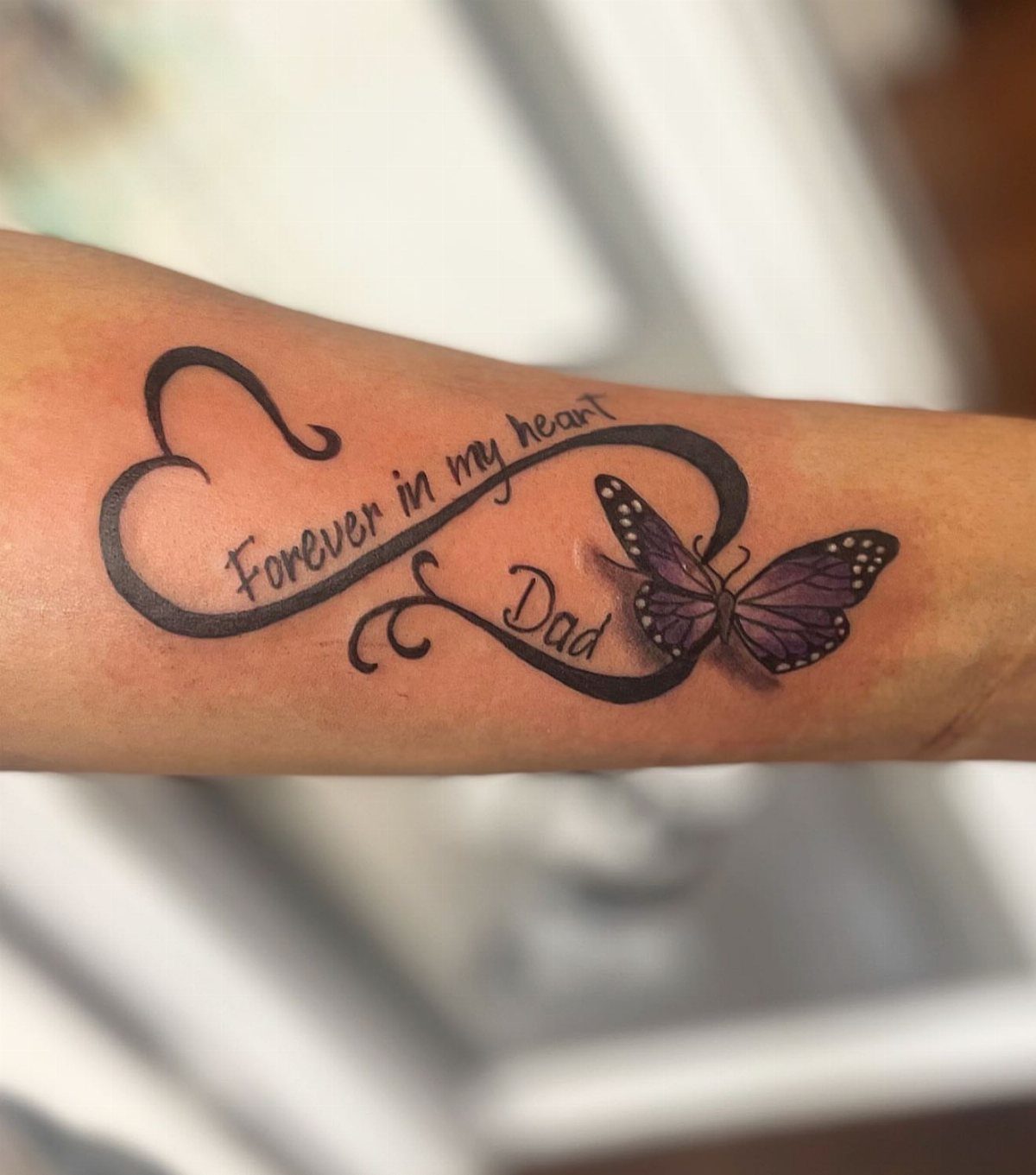 Purple butterfly tattoo with an infinity heart and memorial text on a forearm