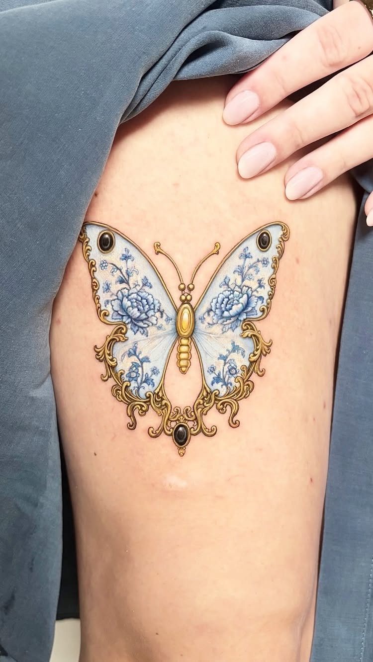 Butterfly tattoo with blue floral patterns and gold ornate edges on a thigh