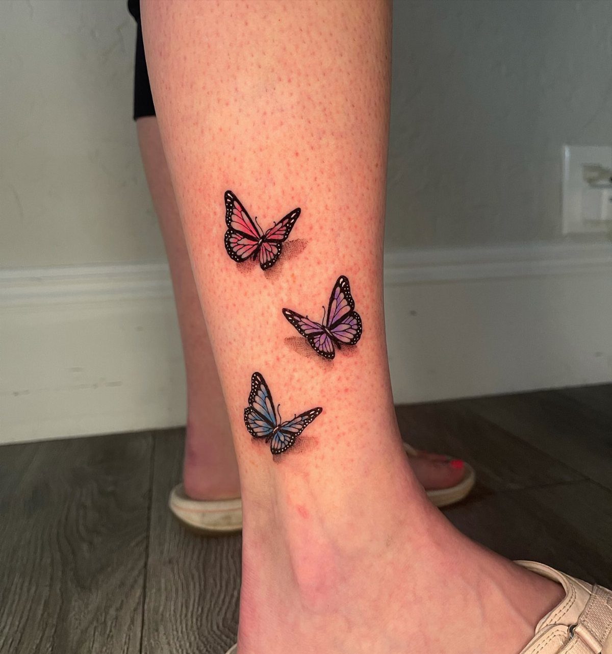Three small blue, pink, and purple butterfly tattoos on a person's ankle.
