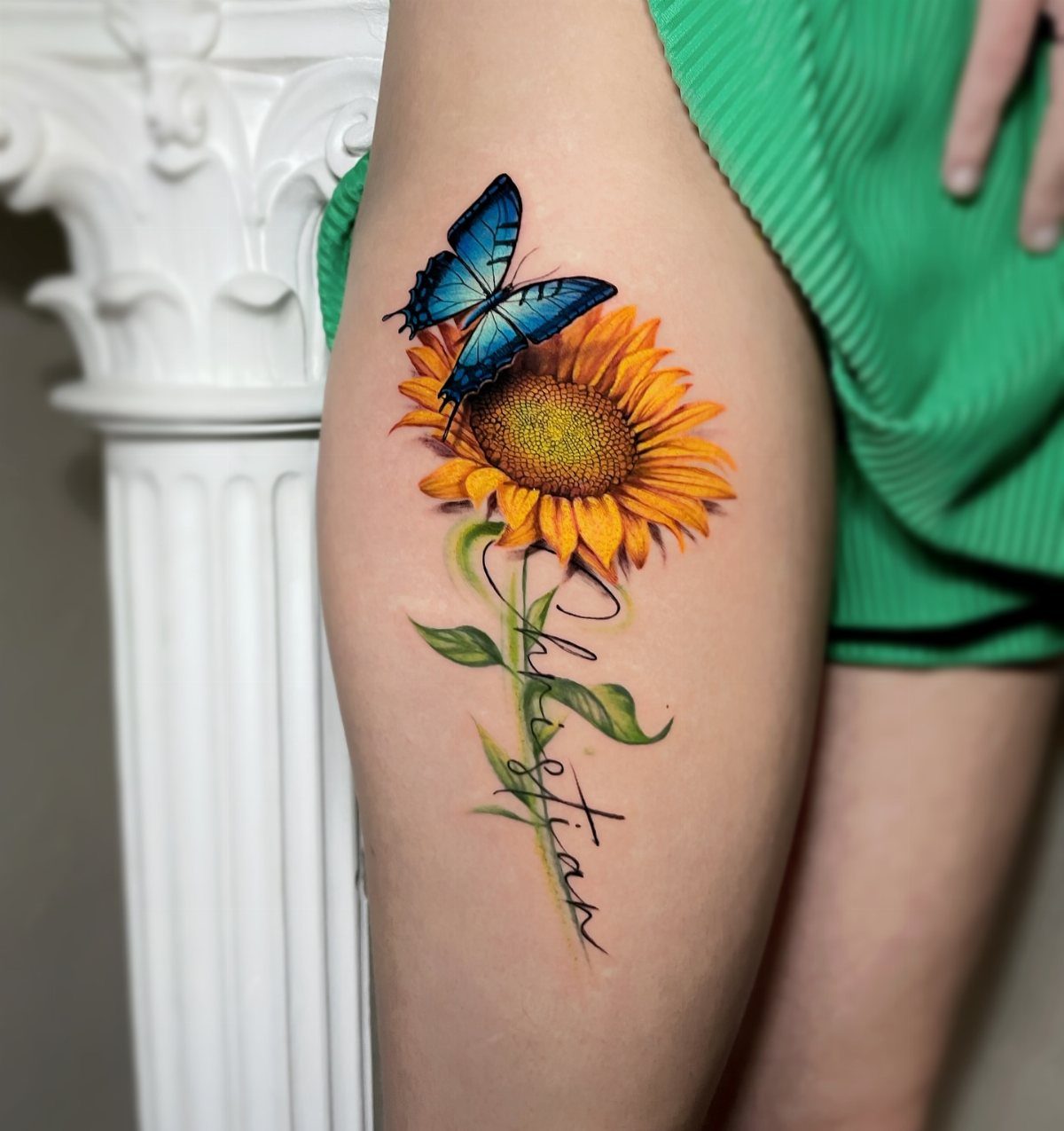 Sunflower and blue butterfly tattoo on a thigh with cursive name text