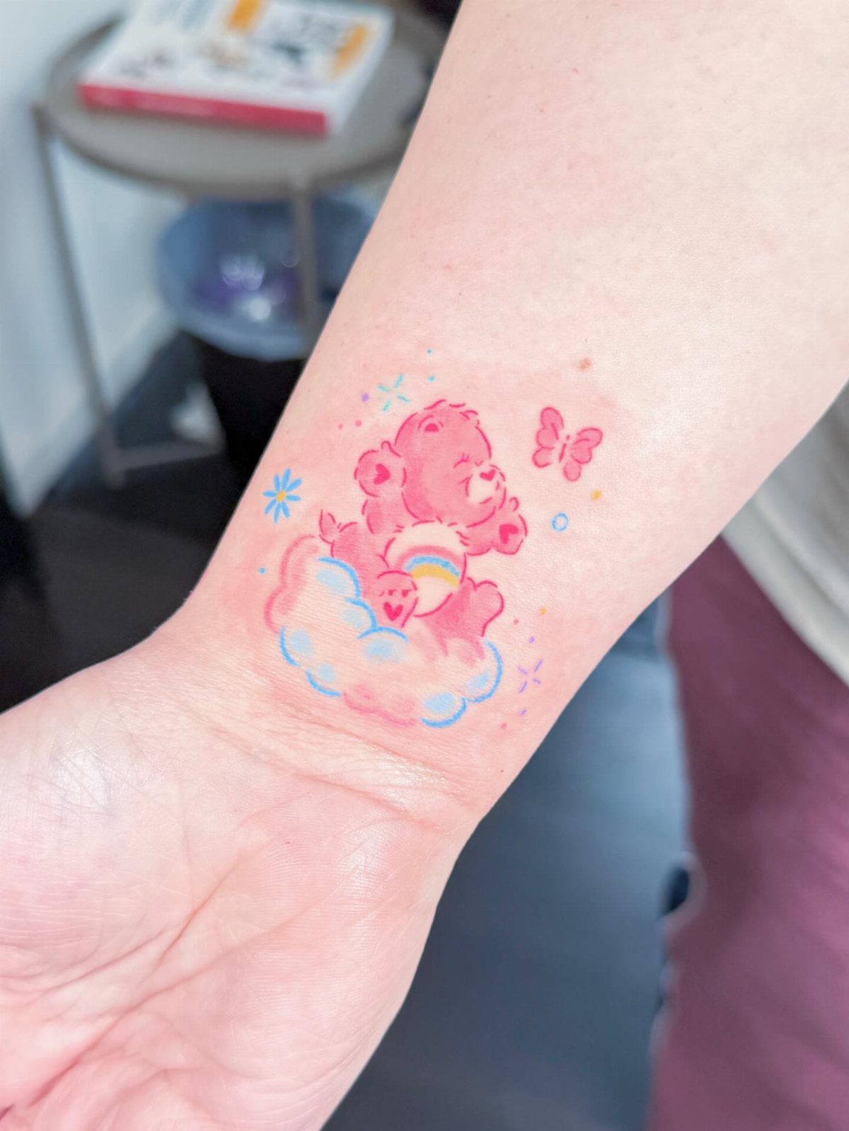 Pink Care Bear tattoo on a wrist with a rainbow, cloud, and butterfly.