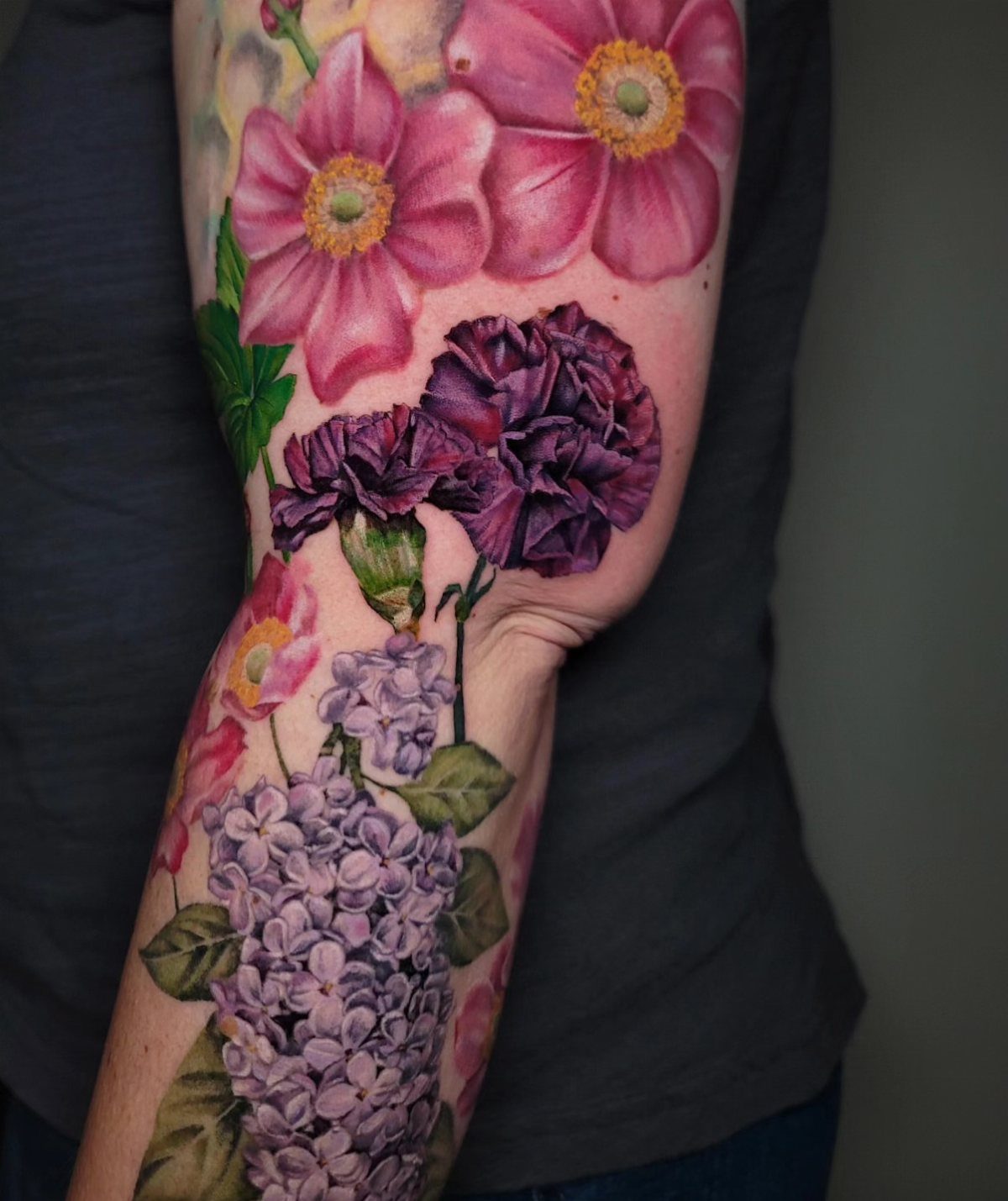 Purple carnation tattoo on an arm surrounded by pink flowers and lilac blossoms
