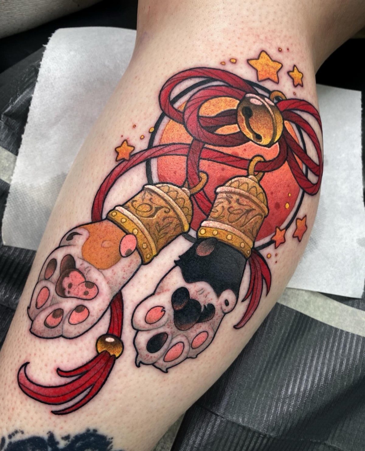 Colorful tattoo of two cat paws with golden bells and red ribbons.
