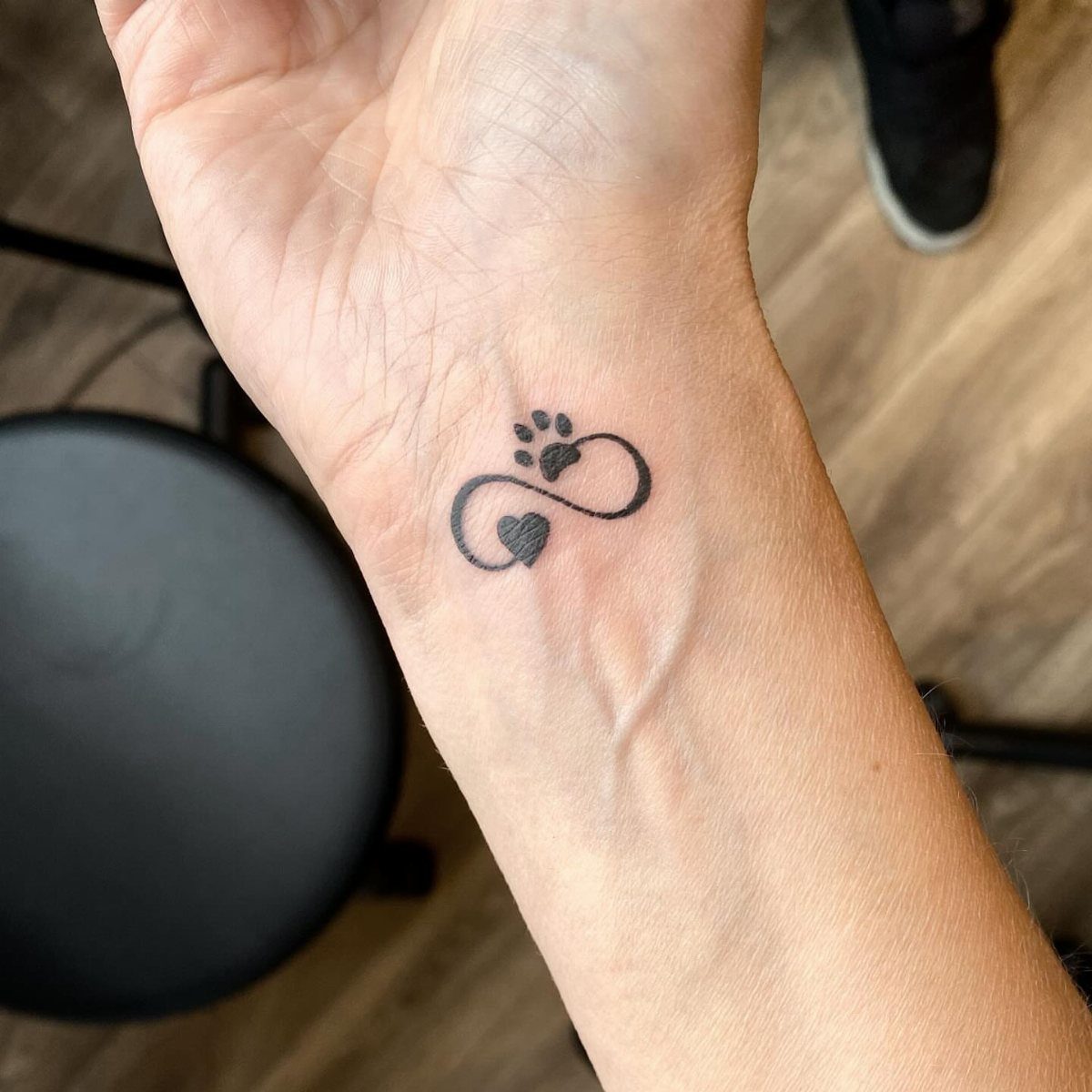 Small black tattoo on a wrist showing a paw print and heart infinity symbol