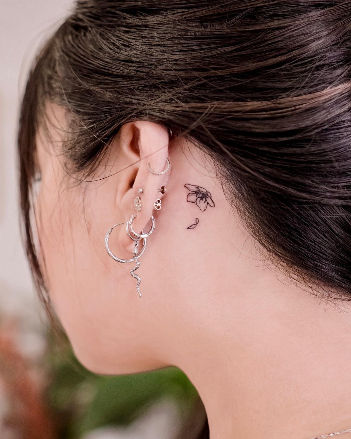 Small black ink fine-line cherry blossom tattoo behind a person's ear with silver earrings