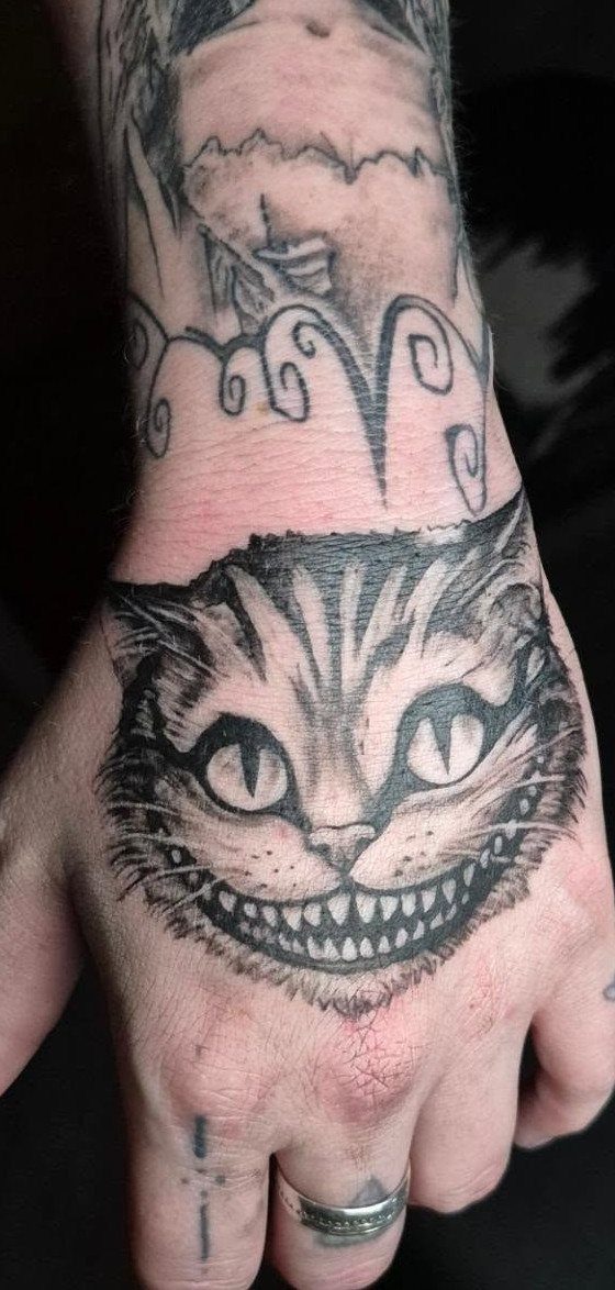 Cheshire Cat tattoo with a wide grin on a person's hand in black ink