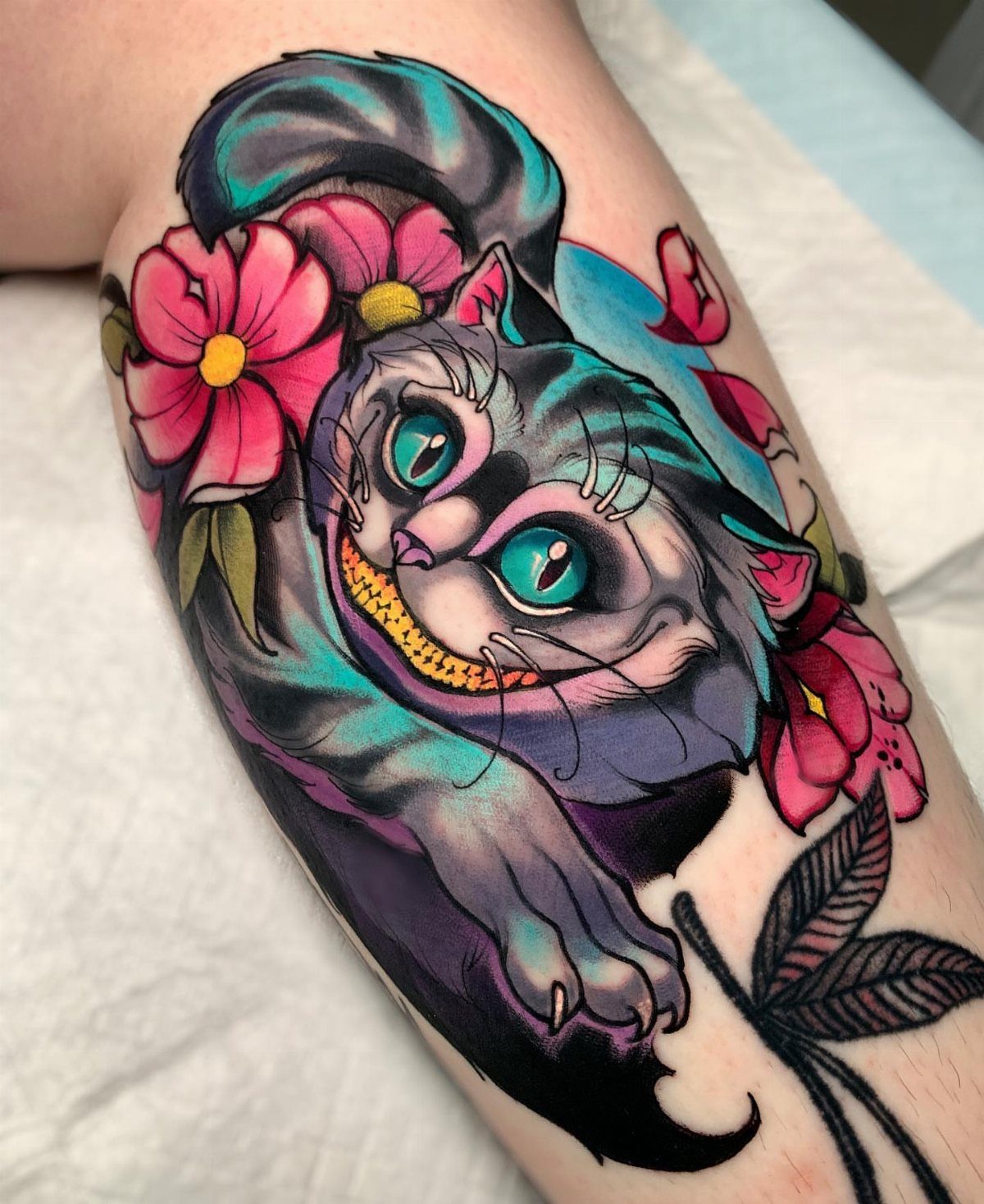 Cheshire Cat tattoo with a wide grin and pink flowers on skin