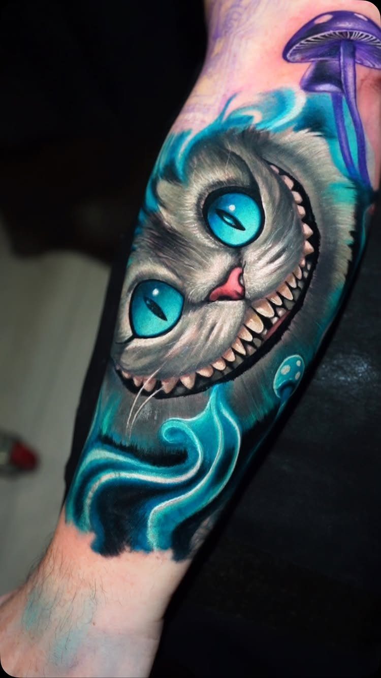 Cheshire Cat tattoo with a wide grin, blue eyes, and a purple mushroom.