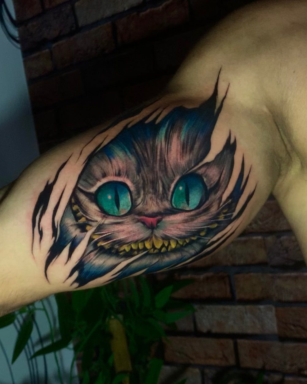 Cheshire Cat tattoo on an arm featuring large teal eyes and a wide grin