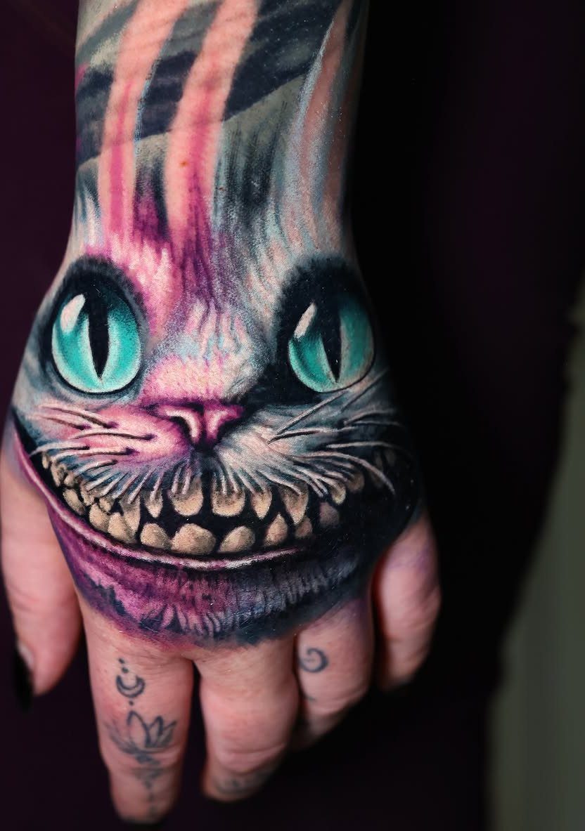 Cheshire Cat tattoo on a hand featuring a wide grin and teal eyes