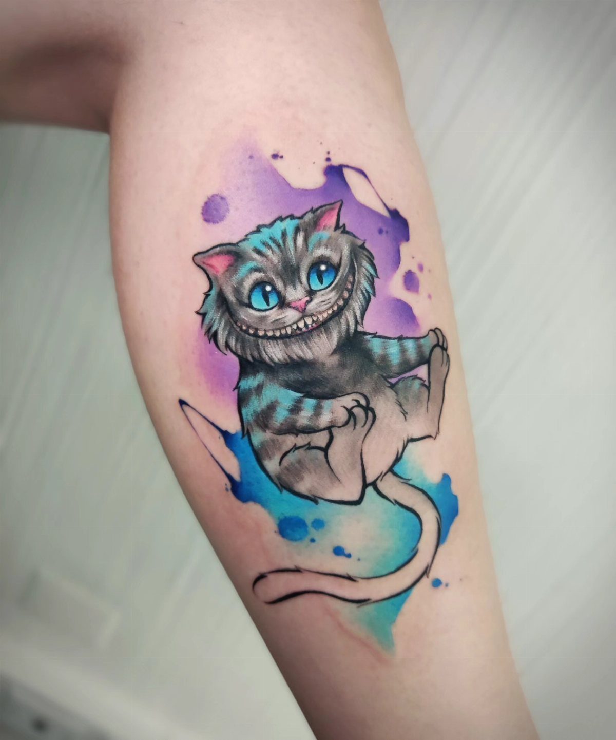 Cheshire Cat tattoo with blue eyes and purple watercolor splashes on a forearm